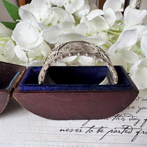 May include: A silver bracelet with an intricate design, resting in a dark brown velvet-lined box. The bracelet has a rounded shape and is decorated with small, raised dots.