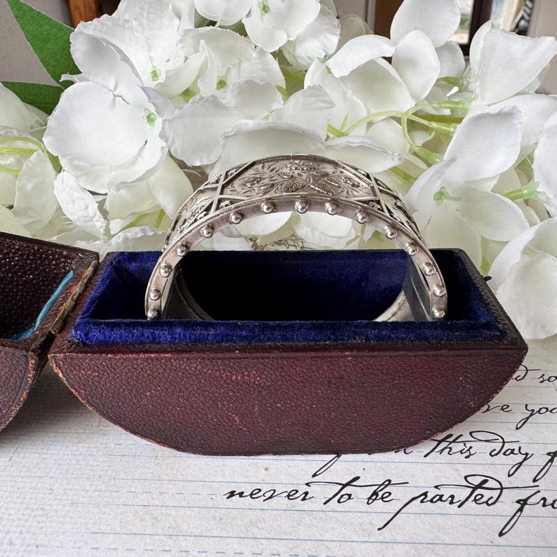 May include: A silver bracelet with an intricate design, resting in a dark brown velvet-lined box. The bracelet has a rounded shape and is decorated with small, raised dots.