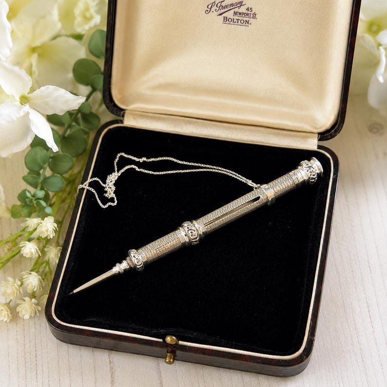 May include: A silver mechanical pencil with an intricate engine-turned pattern and a fine point, resting inside a dark brown hinged presentation box with a black velvet interior and cream silk lining featuring the text J. Freeman 45 Newport St Bolton.