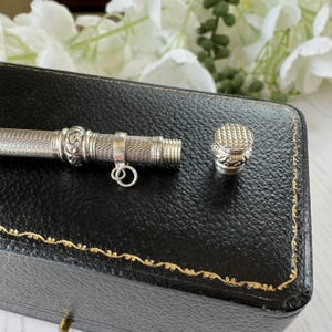 May include: A silver-toned metal cylindrical case with a fine cross-hatched texture, featuring a decorative band with scrollwork, a small attached jump ring, and a matching screw-on cap with a textured top surface and ornate detailing.
