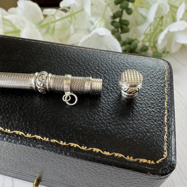May include: A silver-toned metal cylindrical case with a fine cross-hatched texture, featuring a decorative band with scrollwork, a small attached jump ring, and a matching screw-on cap with a textured top surface and ornate detailing.