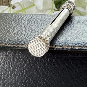 May include: A silver-toned metal mechanical pencil featuring a cylindrical body with a fine cross-hatched knurled texture. The top end has a circular, flat cap with a diamond-patterned grip, and the barrel includes a thin, vertical slot.