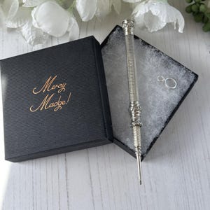 May include: A silver-toned metal stylus with a textured, ribbed barrel and decorative filigree accents, presented in a black square gift box with the text Mercy Madge! in gold script on the lid, accompanied by a small silver jump ring.