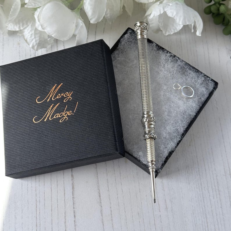 May include: A silver-toned metal stylus with a textured, ribbed barrel and decorative filigree accents, presented in a black square gift box with the text Mercy Madge! in gold script on the lid, accompanied by a small silver jump ring.
