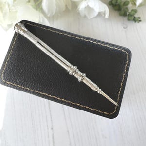 May include: A silver mechanical pencil featuring a textured, cross-hatched metal barrel with decorative bands and a pointed tip. The writing instrument has a slim, cylindrical body with intricate detailing along the shaft and a small side slot.