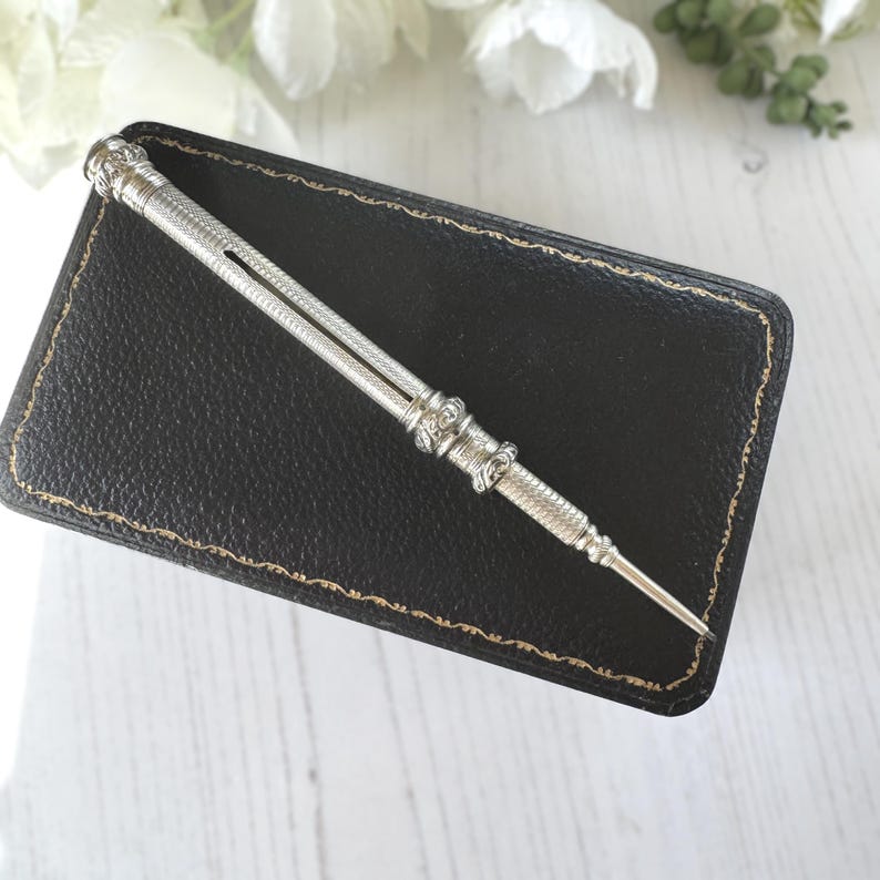 May include: A silver mechanical pencil featuring a textured, cross-hatched metal barrel with decorative bands and a pointed tip. The writing instrument has a slim, cylindrical body with intricate detailing along the shaft and a small side slot.