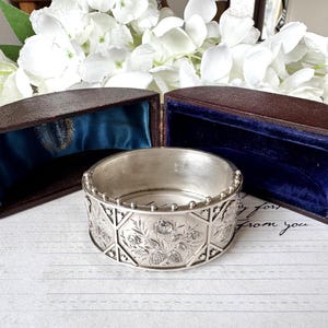 May include: A silver bangle bracelet with an intricate floral design. The bracelet is in a brown box with a blue velvet lining.