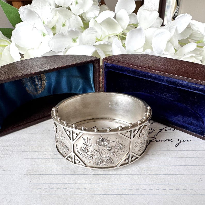 May include: A silver bangle bracelet with an intricate floral design. The bracelet is in a brown box with a blue velvet lining.
