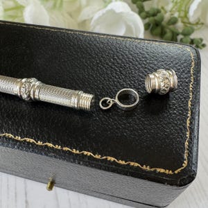 May include: A silver-toned cylindrical metal case with a textured cross-hatch pattern and ornate floral bands. The piece includes a detachable screw-top cap and a small circular metal ring attachment, presented as a disassembled jewelry component.