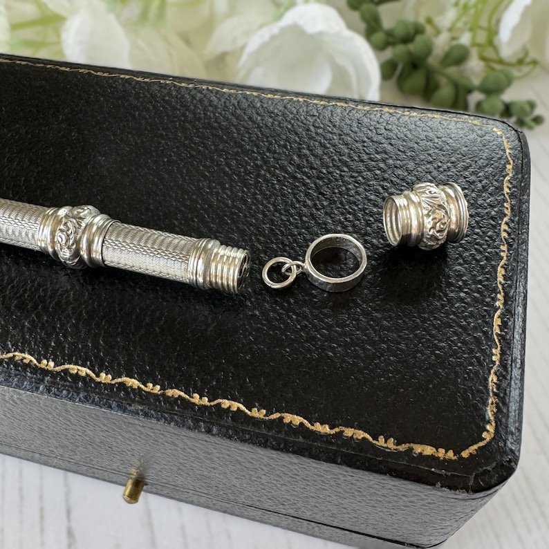 May include: A silver-toned cylindrical metal case with a textured cross-hatch pattern and ornate floral bands. The piece includes a detachable screw-top cap and a small circular metal ring attachment, presented as a disassembled jewelry component.