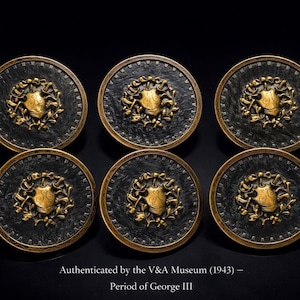 Peut inclure: Six objets décoratifs antiques circulaires, avec un fond noir et des détails dorés. Chacun présente un motif central de bouclier doré entouré d'une couronne de laurier dorée. Le texte en bas indique "Authenticated by the V&A Museum (1943) - Period of George III".