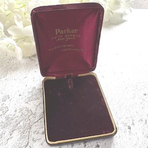 May include: An open, burgundy velvet jewelry box with gold trim. The interior features a plush lining and the name "Parker Fifth Avenue New York" printed in gold lettering. The box is designed to hold a necklace or pendant.