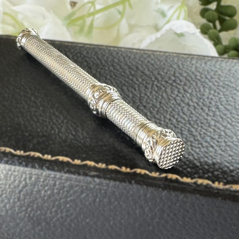 May include: A cylindrical silver needle case featuring a fine cross-hatched texture along its length, accented by decorative embossed bands and a flat, grid-patterned cap at one end. The metal surface reflects light, highlighting the intricate detailing.