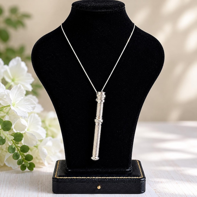 May include: A silver-toned cylindrical pendant suspended from a thin silver chain. The pendant features a textured, cross-hatched surface with decorative bands at the top, middle, and bottom, displaying a metallic finish and a slender, elongated form.