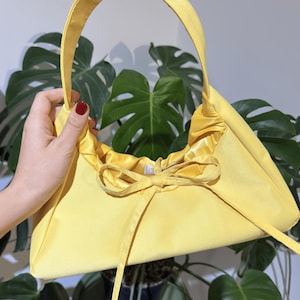 May include: A vibrant yellow handbag with a curved handle. The bag features a drawstring closure with a bow detail. The bag is made of a soft, satin-like material. Large green leaves form the background.