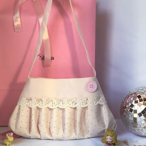 Peut inclure: Un sac à main rose clair doté d'une section inférieure plissée avec de petits pois noirs, d'une bordure en dentelle blanche le long du bord supérieur et d'un grand bouton rose. Le sac possède une longue bandoulière en tissu lisse.