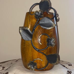 Può includere: Un vaso di legno con finitura marrone scuro e un tralcio decorativo in metallo con foglie e fiori. I dettagli in metallo hanno una finitura nera e oro, aggiungendo un tocco di eleganza al vaso. Un elemento decorativo per la casa.
