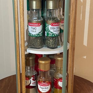 May include: A wooden spice rack with three tiers, holding several glass spice jars with gold lids. The rack has a natural wood finish with green accents. The jars contain various herbs and spices.