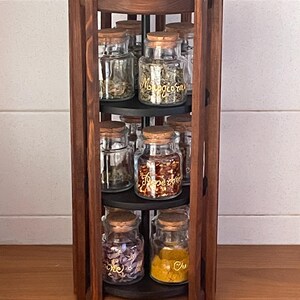Peut inclure: Un présentoir à épices en bois brun foncé avec plusieurs étagères, contenant des bocaux en verre transparent remplis de diverses épices. Les bocaux ont des couvercles en liège et des étiquettes dorées. Le présentoir pivote pour un accès facile aux épices.