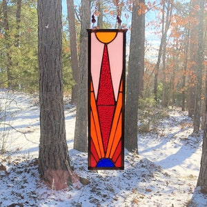 May include: A stained glass suncatcher with a geometric design in shades of pink, red, orange, yellow, and blue. The suncatcher is hanging from a chain in front of a window with a snowy forest scene.