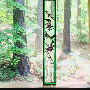 May include: A stained glass window panel with a green border and a design of three purple flowers with green leaves and a hummingbird in flight.