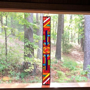 May include: A stained glass window hanging in a window frame. The window is made of colorful glass pieces in red, orange, yellow, green, blue, and white. The window is a geometric design with a variety of shapes.