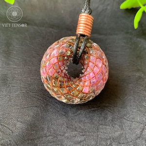 Pode incluir: Um colar com pingente com um design esférico de centro aberto. O pingente é feito com pedras rosa e transparentes, envoltas em uma grade de fio de cobre. O colar tem um cordão preto e um detalhe em cobre na parte superior.