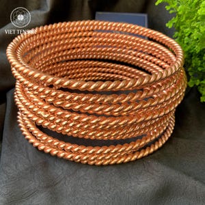 May include: A collection of copper-toned bangles featuring a twisted rope design. The bangles are stacked, showcasing a textured surface created by the rope pattern. The jewellery is presented on a dark background.