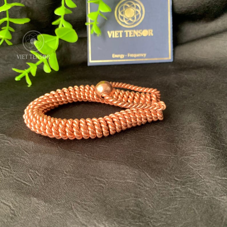 May include: A copper-colored bracelet with a woven design and a spherical bead. The bracelet is made of tightly wound wire and is displayed on a dark surface. A tag with the text "VIET TENSOR" is in the background.