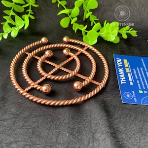 Könnte beinhalten: Ein kupferfarbener Metall-Topfuntersetzer mit einem gedrehten Seildesign. Der Topfuntersetzer hat eine kreisförmige Form mit einem zentralen Design und kleinen Kugelakzenten. Eine blaue Karte mit dem Text "THANK YOU" ist sichtbar.