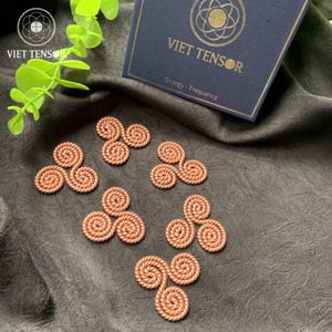 Puede incluir: Dispositivos energéticos en espiral de color cobre dispuestos sobre una superficie gris oscuro. Los dispositivos tienen un diseño de tres pétalos. Una caja azul marino con texto dorado "VIET TENSOR" está en el fondo. La caja también tiene el texto "Energy - Frequency".
