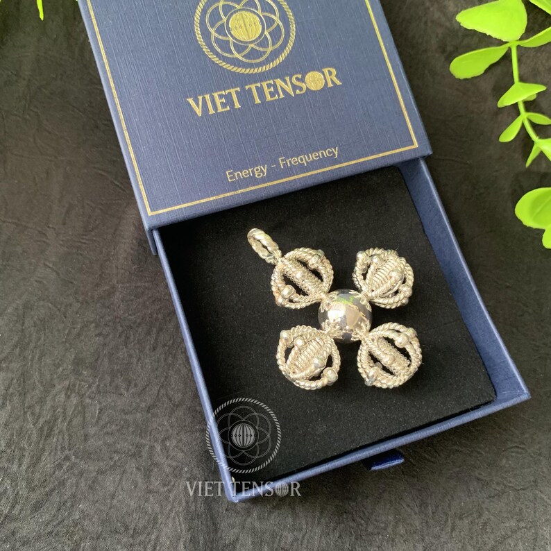 May include: A silver pendant in a cross shape with a central clear stone, presented in a blue box. The box is labeled "VIET TENSOR" and "Energy - Frequency". The pendant has an intricate, woven design.