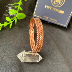 May include: A copper-colored bracelet with a braided design rests on a clear crystal point. The bracelet is displayed with a box that reads "VIET TENSOR" and "Energy - Frequency". Green leaves are visible in the background.