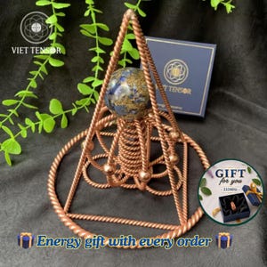 Könnte beinhalten: Eine Pyramidenstruktur aus Kupferdraht mit einer zentralen Spiralfederbasis, die von einer blau-goldenen Kugel gekrönt wird. Der Rahmen besteht aus gedrehtem Kupferdraht, einschließlich einer kreisförmigen Basis. Eine dunkelblaue Box mit dem Text VIET TENSOR ist sichtbar.