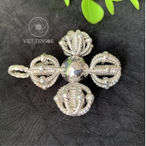 May include: A silver-colored pendant with a cross-like design. The pendant features a central sphere and four ornate, looped arms. The text "VIET TENSOR" is visible in the upper left corner. The pendant is displayed on a black surface.