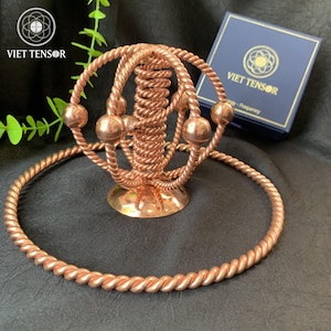 Könnte beinhalten: Ein kupferfarbener Tensorring und eine passende Halskette liegen auf einer dunklen Oberfläche. Der Tensorring hat ein kugelförmiges Design mit vier Kugeln und den Text "VIET TENSOR" auf einer blauen Box im Hintergrund.