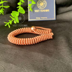 May include: A copper-colored bracelet with a woven design and a spherical bead. The bracelet is displayed on a dark surface, with a card that reads "VIET TENSOR" and "Energy - Frequency".