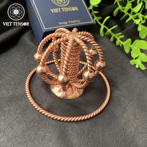 Può includere: Un tensore in metallo color rame con un design sferico, caratterizzato da una colonna centrale e anelli circostanti con sfere. Il tensore è esposto su una superficie scura, con una scatola con l'etichetta "VIET TENSOR" sullo sfondo.