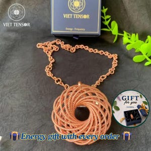 Op de afbeelding: Een koperkleurige hangerketting met een cirkelvormig, spiraalvormig draadontwerp dat aan een bijpassende ketting hangt. De hanger wordt getoond naast een donkerblauwe doos met het label VIET TENSOR en een cirkelvormige afbeelding van een geschenkdoos met de tekst GIFT for you 333MHZ.