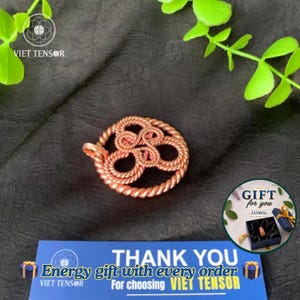 Op de afbeelding: Een ronde hanger gemaakt van gedraaid koperdraad in een ingewikkeld knooppatroon. De hanger wordt getoond naast een blauwe kaart met de tekst THANK YOU Energy gift with every order VIET TENSOR en een rond cadeaukaartje met tekst.