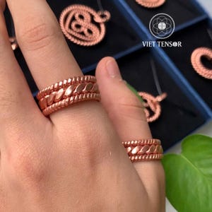 Op de afbeelding: Close-up van koperkleurige ringen met een gevlochten ontwerp op een hand. De ringen hebben een getextureerd uiterlijk en zijn waarschijnlijk gemaakt van koper. Andere koperkleurige sieraden zijn zichtbaar op de achtergrond, samen met de tekst "VIET TENSOR".