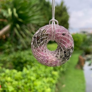 Puede incluir: Un colgante circular con un diseño de alambre que envuelve pequeñas piedras pulidas de color rosa y gris metálico. El colgante está suspendido de una fina cadena de color plateado, creando un adorno geométrico en forma de rosquilla.