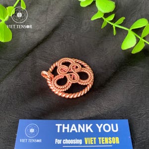 Może przedstawiać: Miedziany wisiorek o okrągłym wzorze, z plecionym wzorem przypominającym linę. Wisiorek ma pętlę na łańcuszek i jest umieszczony na ciemnym tle z zielonymi liśćmi. Widoczna jest również niebieska karta z napisem "THANK YOU".