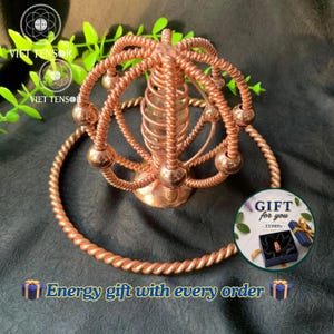 Puede incluir: Una escultura de alambre color cobre con una bobina vertical central rodeada por una jaula esférica de alambre trenzado y pequeñas esferas, sobre una base circular. Un recuadro circular muestra una caja de regalo con un anillo y texto.