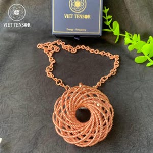 May include: A copper-colored pendant necklace with a circular, woven design. The pendant features a central hole and is connected to a chain necklace. The image also shows a box with the text "VIET TENSOR" and "Energy - Frequency".