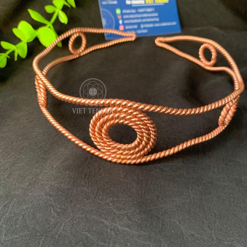 May include: A copper-colored metal headband constructed from twisted wire. The design features a continuous rope-like band accented with three prominent circular motifs, each formed by coiled wire, positioned at the front and sides.