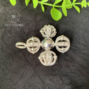 May include: A silver-colored, ornate pendant with a central sphere and four decorative elements. Each element features a looped design with intricate detailing. The pendant is set against a dark background with green foliage.