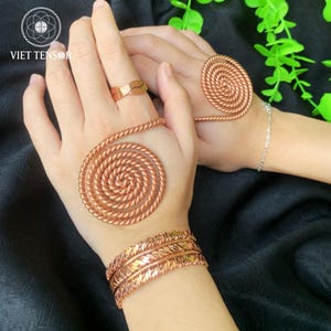 Puede incluir: Juego de joyas de cobre con un anillo, adornos de mano en espiral y una pulsera. El anillo tiene un diseño de onda único. Las piezas en espiral están hechas de alambre de cobre enrollado. La pulsera tiene un patrón de hojas texturizadas. Las joyas se exhiben sobre un fondo oscuro.