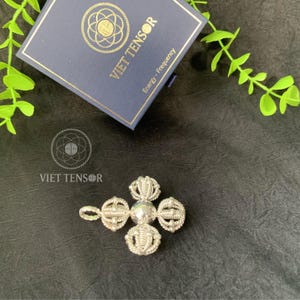 May include: A silver-colored pendant with a central sphere and four ornate, circular designs. The pendant is next to a navy blue box with gold lettering that reads "VIET TENSOR" and "Energy - Frequency". The image is on a dark surface.