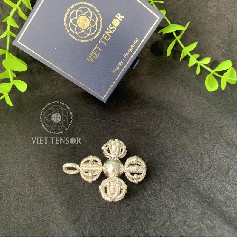 May include: A silver-colored pendant with a central sphere and four ornate, circular designs. The pendant is next to a navy blue box with gold lettering that reads "VIET TENSOR" and "Energy - Frequency". The image is on a dark surface.